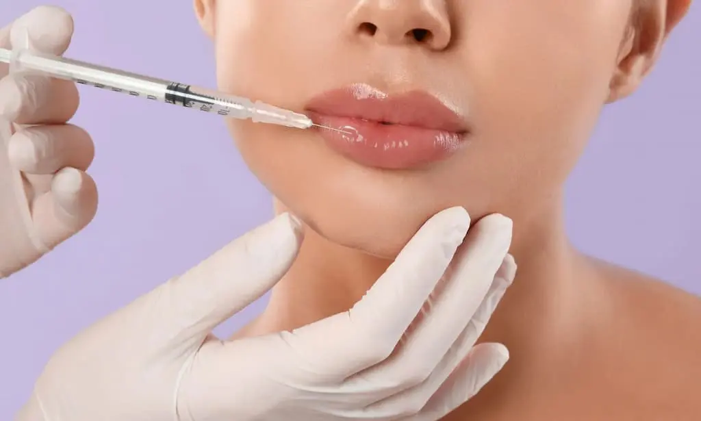 A person receives a lip injection from a gloved medical professional against a light purple background.