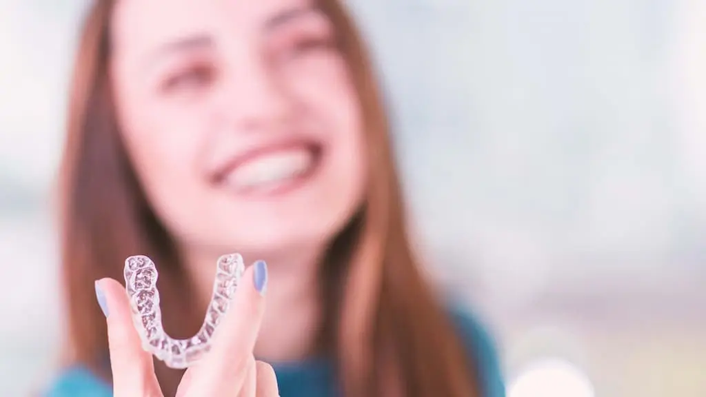 A person holds a clear dental aligner with their right hand, smiling in the background.