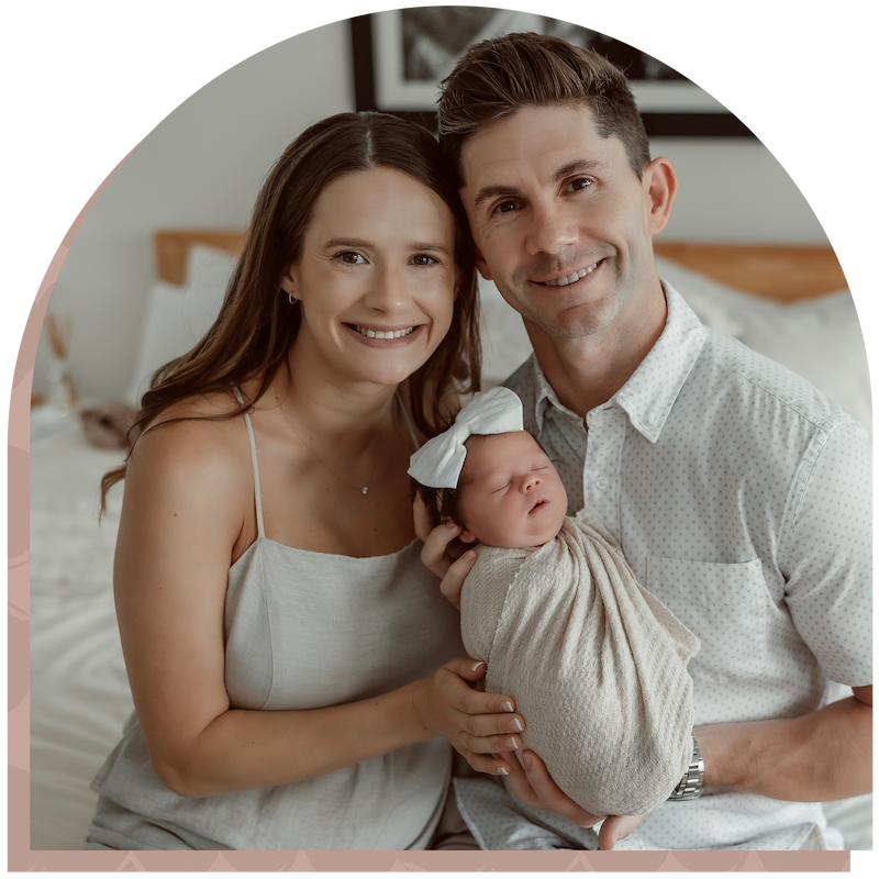 A smiling couple sits closely together on a bed, holding a swaddled newborn baby with a bow headband.