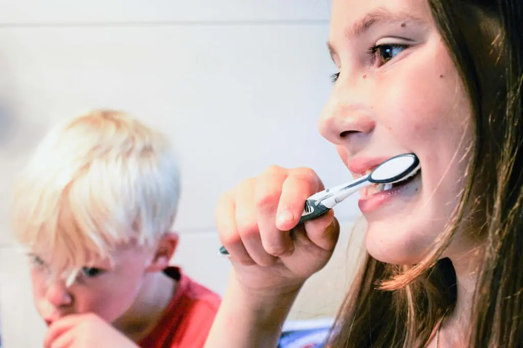 Two children brush their teeth; a girl is in the foreground smiling with a toothbrush, while a boy in the background focuses on brushing.