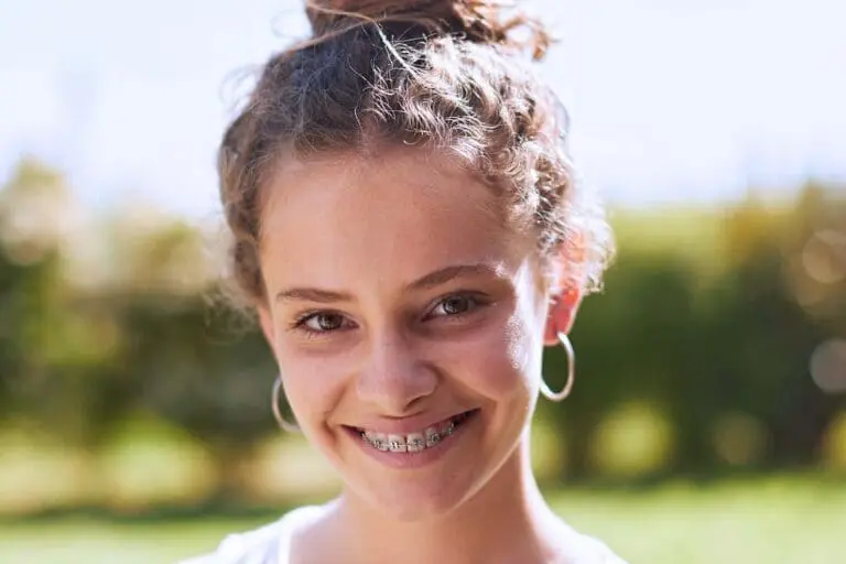 Close-up of a smiling young person with curly hair in a bun, wearing hoop earrings and braces, outdoors with a blurred green background.