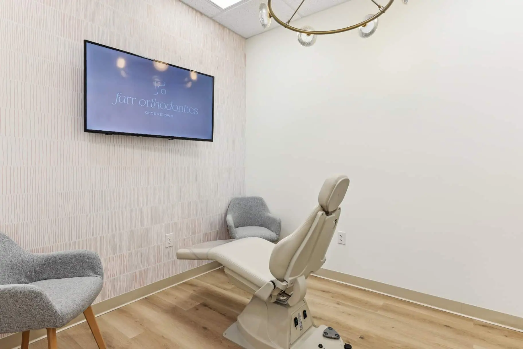 A modern orthodontic exam room with a reclining chair, two gray armchairs, a TV displaying "Farr Orthodontics Georgetown," and light wood flooring at Farr Orthodontics Georgetown in Georgetown, TX.