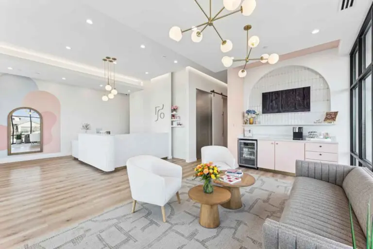 Modern waiting room with a gray sofa, white chairs, coffee tables, fresh flowers, magazines, a TV, mini-fridge, and a white reception desk, featuring large windows and stylish lighting at Farr Orthodontics Georgetown, Georgetown, TX.