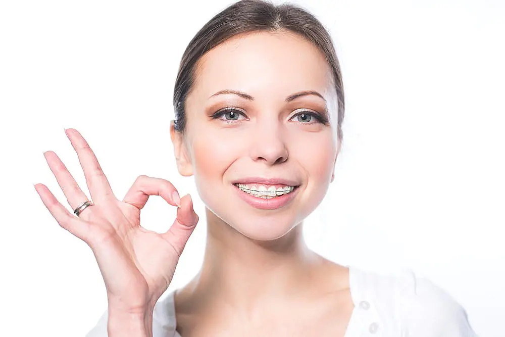 A young woman with clear ceramic braces smiles and makes an "OK" hand gesture against a white background at Farr Orthodontics in Georgetown, TX.