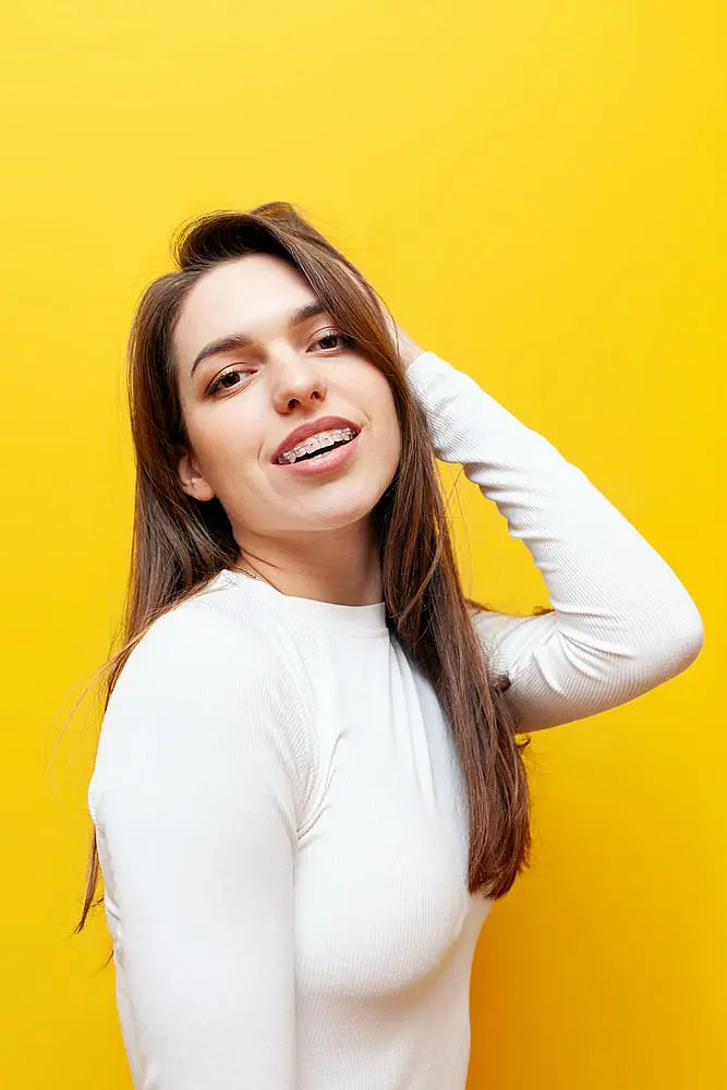 A young woman with long brown hair, clear ceramic braces, and a white long-sleeve shirt smiles against a bright yellow background at Farr Orthodontics in Georgetown, TX.