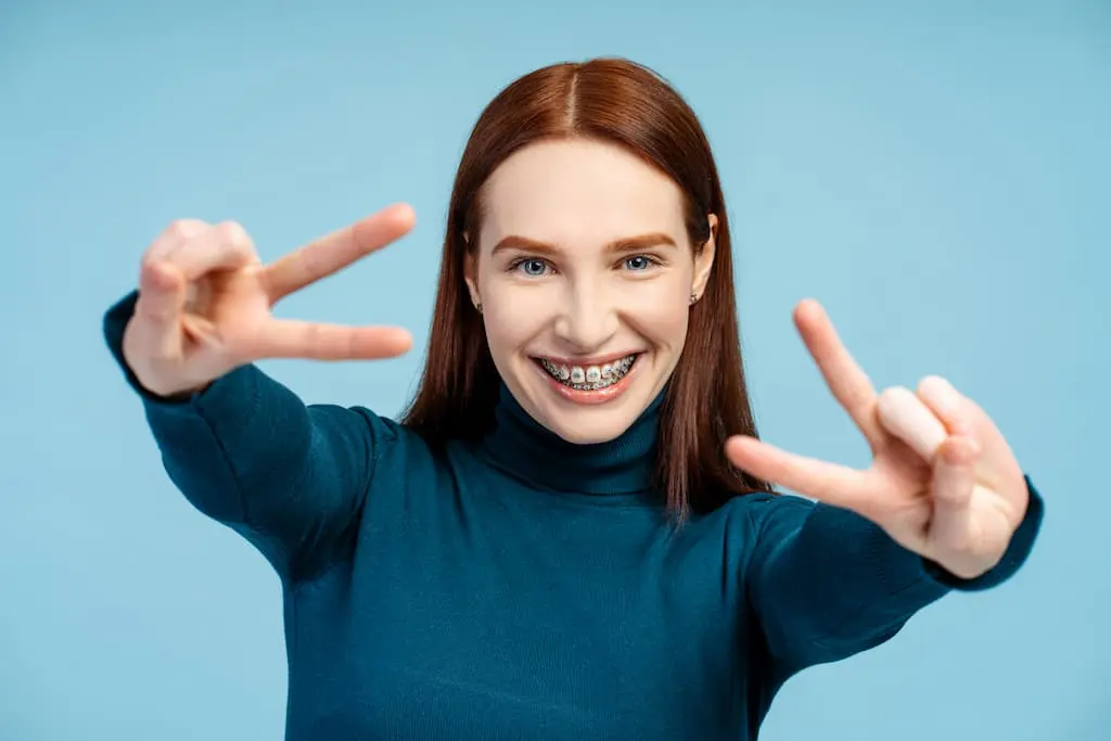 Young woman with long brown hair, wearing a teal turtleneck and braces, smiles and makes peace signs with both hands against a plain blue background in Georgetown, TX.