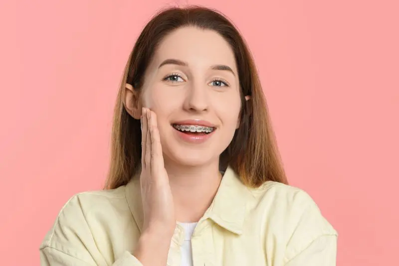 Young woman with long brown hair and braces smiling, holding her hand to her cheek, standing in front of a pink background, wearing metal braces in Georgetown TX.