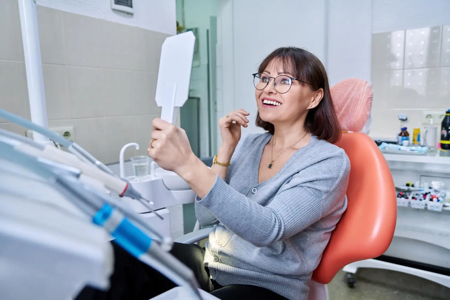 A woman smiles at her reflection in a hand mirror at Farr Orthodontics in Georgetown, TX, surrounded by orthodontic appliances.