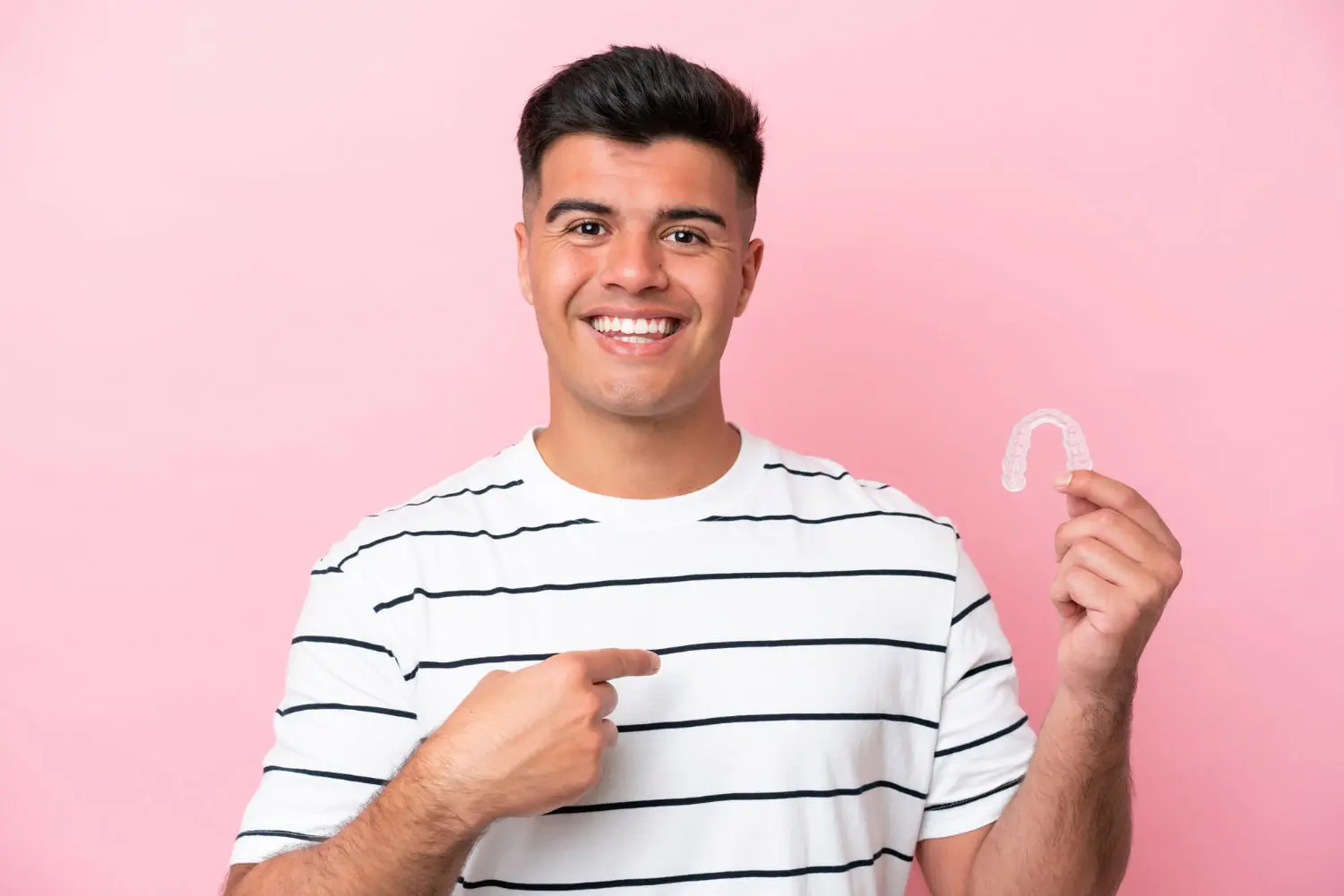 A man in a striped shirt holds and points to an Invisalign aligner, smiling. Represent Farr Orthodontics in Georgetown, TX pink backdrop.