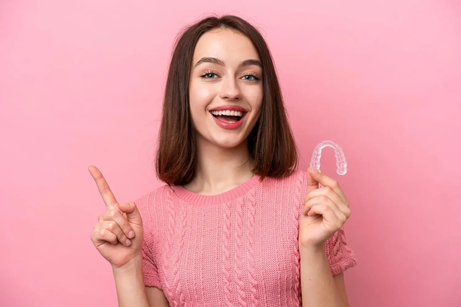 Smiling young woman in a pink sweater holds an Invisalign aligner at Farr Orthodontics in Georgetown, TX, with pink background.