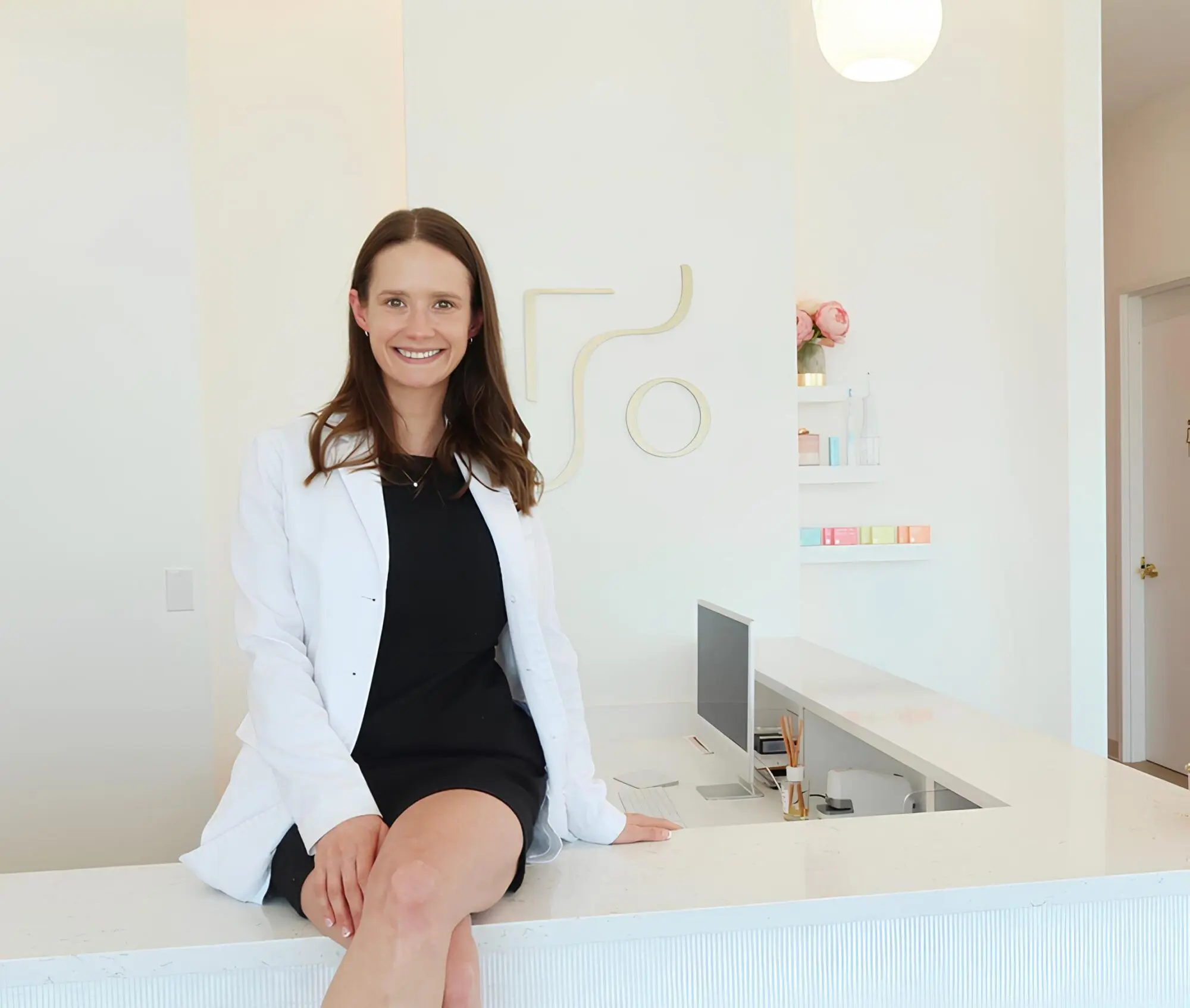 A woman in a white coat sits on the edge of a white reception desk in a bright, modern office representing all PPO insurance plans accepted at Farr Orthodontics Georgetown in Georgetown, TX.