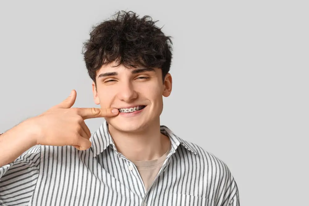 Smiling young man points to his braces, sharing about braces cost at Farr Orthodontics in Georgetown, TX, on a gray background.