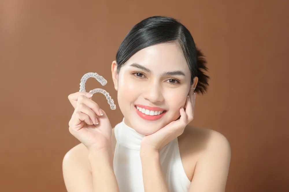A woman smiles, holding clear dental aligners; Farr Orthodontics in Georgetown, TX features Invisalign cost options with a brown backdrop.