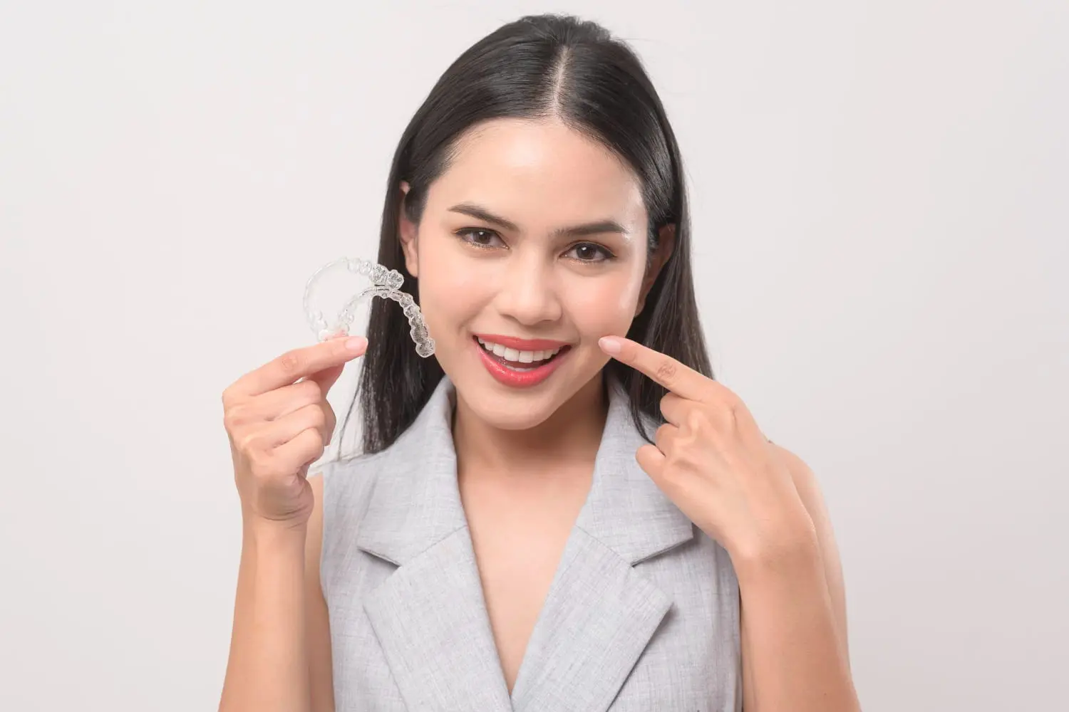 At Farr Orthodontics in Georgetown, TX, a woman in a gray sleeveless blazer smiles while holding an Invisalign aligner and pointing to her teeth, showing her excitement about affordable Invisalign treatment.