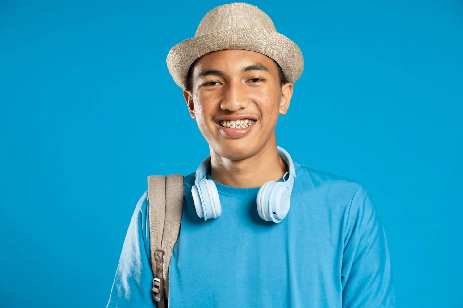 Smiling youth with affordable braces from Farr Orthodontics in Georgetown, TX, wearing hat, blue shirt, headphones, backpack on blue background.