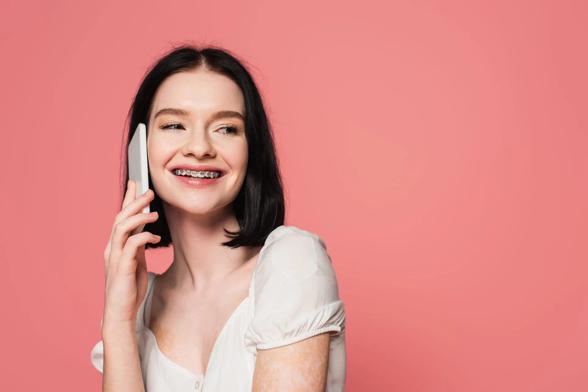 Smiling with affordable braces from Farr Orthodontics in Georgetown, TX, a young woman talks on her smartphone against a pink background.